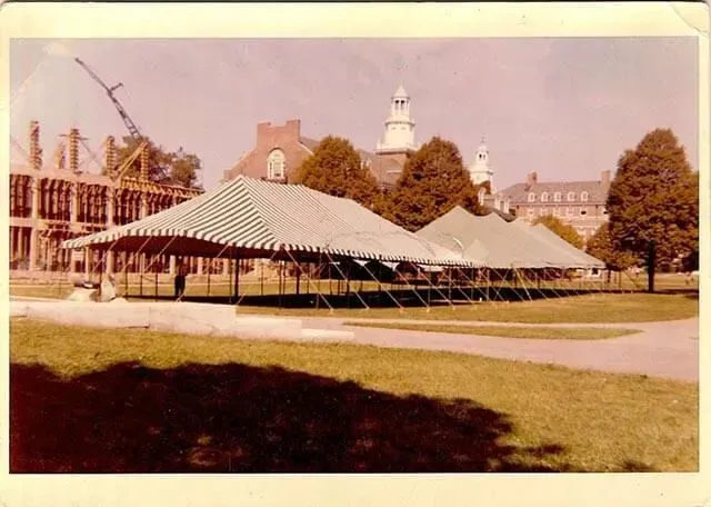 A large, striped event tent set up on a university lawn in front of brick buildings under a clear sky.