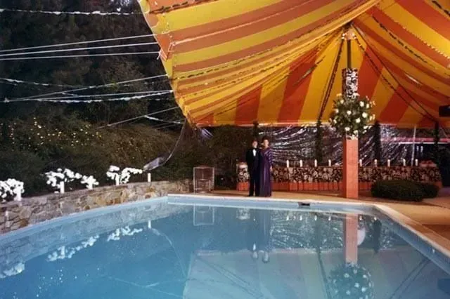A couple stands under a yellow and orange striped canopy next to a pool at an outdoor event.