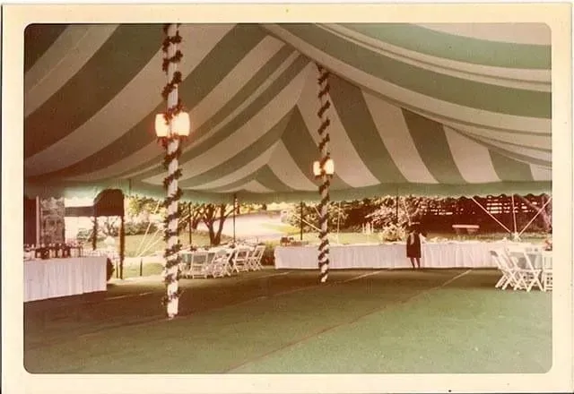 An event tent with green and white striped fabric, decorated poles, white tables, and chairs on a green floor.