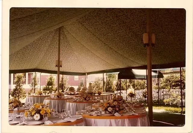Outdoor tent set up for a celebration with round tables decorated with yellow flowers, white linens, and chairs.