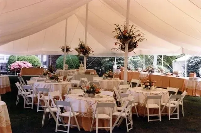 White tent set up for an event, featuring round tables with white tablecloths, chairs, and floral centerpieces.