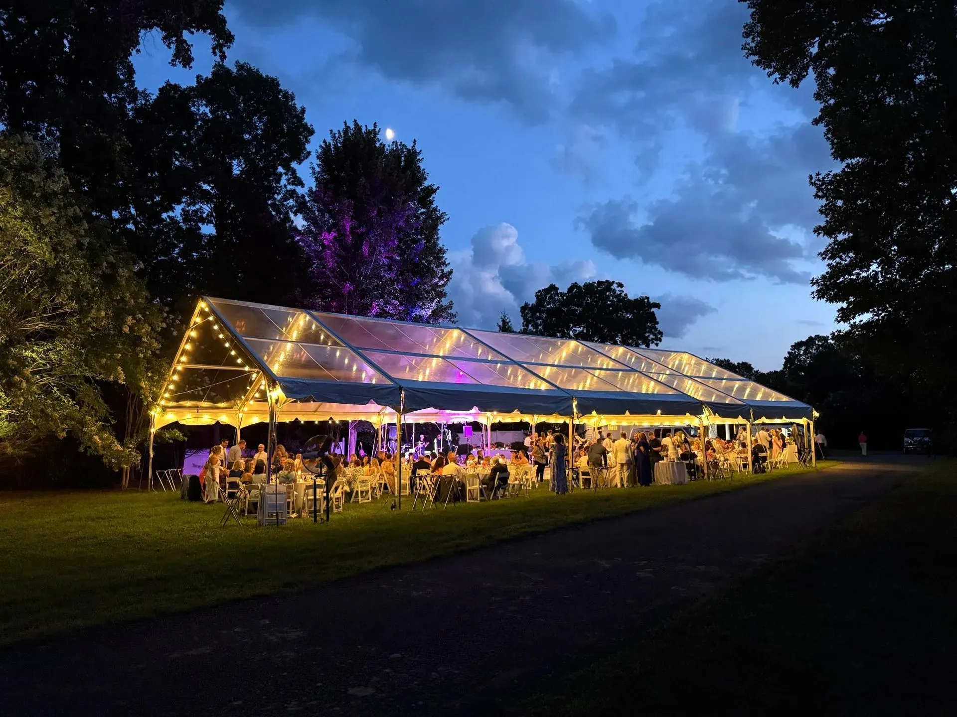 A clear event tent illuminated with warm string lights at dusk, set on a lawn with guests seated at tables inside.