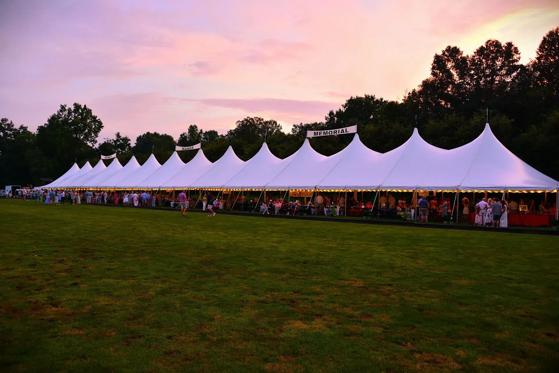 A long white tent with many peaks sits on a grassy field under a vibrant purple and orange sunset.