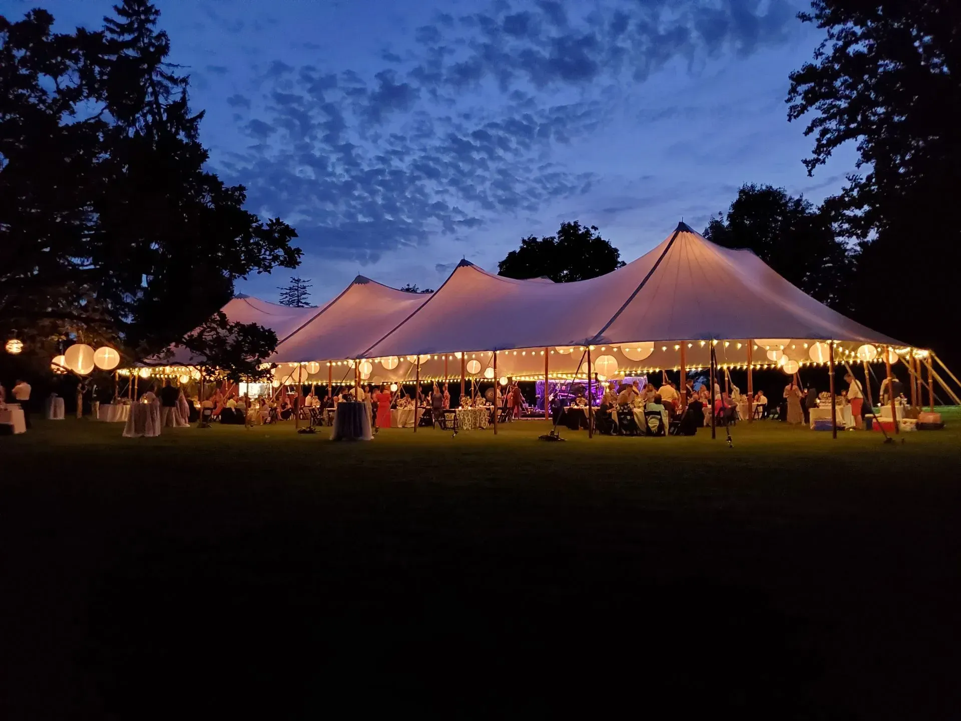 A large, illuminated white event tent glows on a green lawn at twilight, surrounded by trees and string lights.