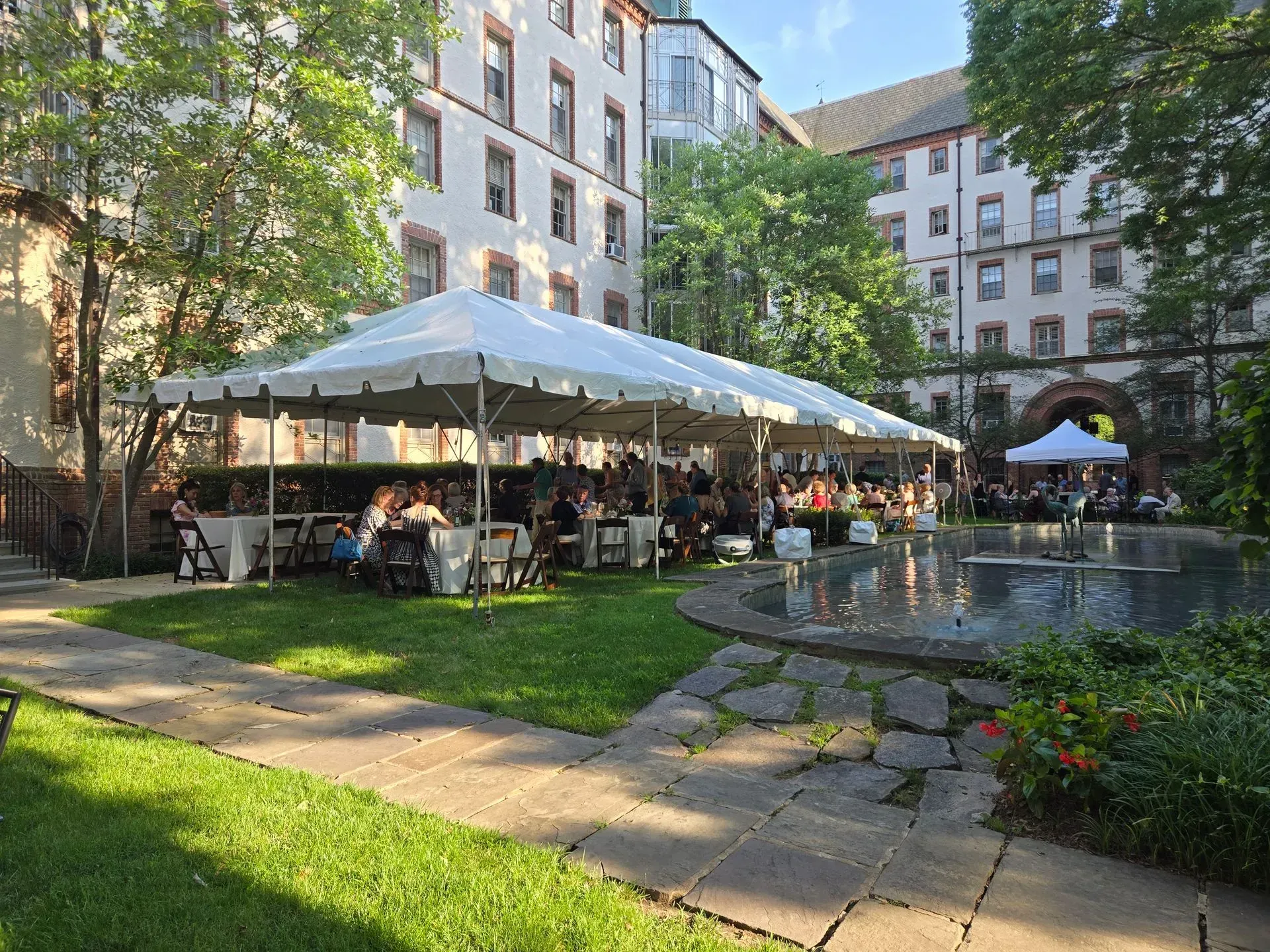 A long white event tent set up in a courtyard with tables, chairs, and guests near a stone fountain and multi-story building.