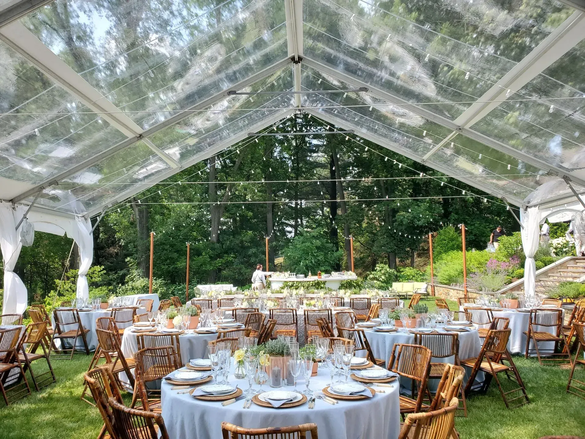 An outdoor wedding tent with a clear roof set up for a reception with round tables, white linens, and wooden chairs.