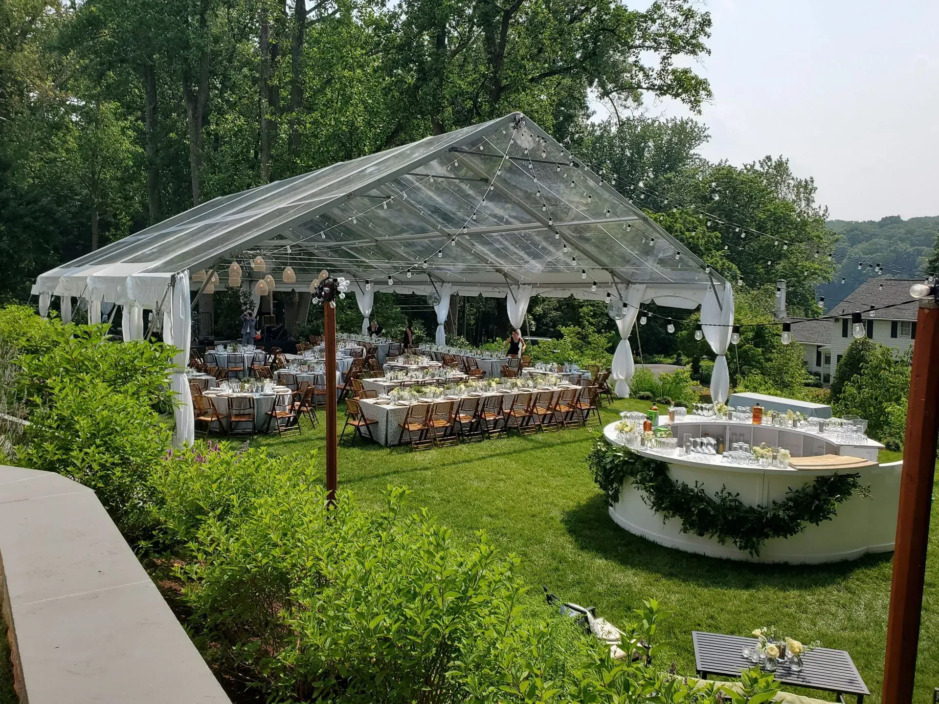 A clear-topped outdoor event tent covers dining tables on a green lawn, with a circular bar decorated with greenery nearby.