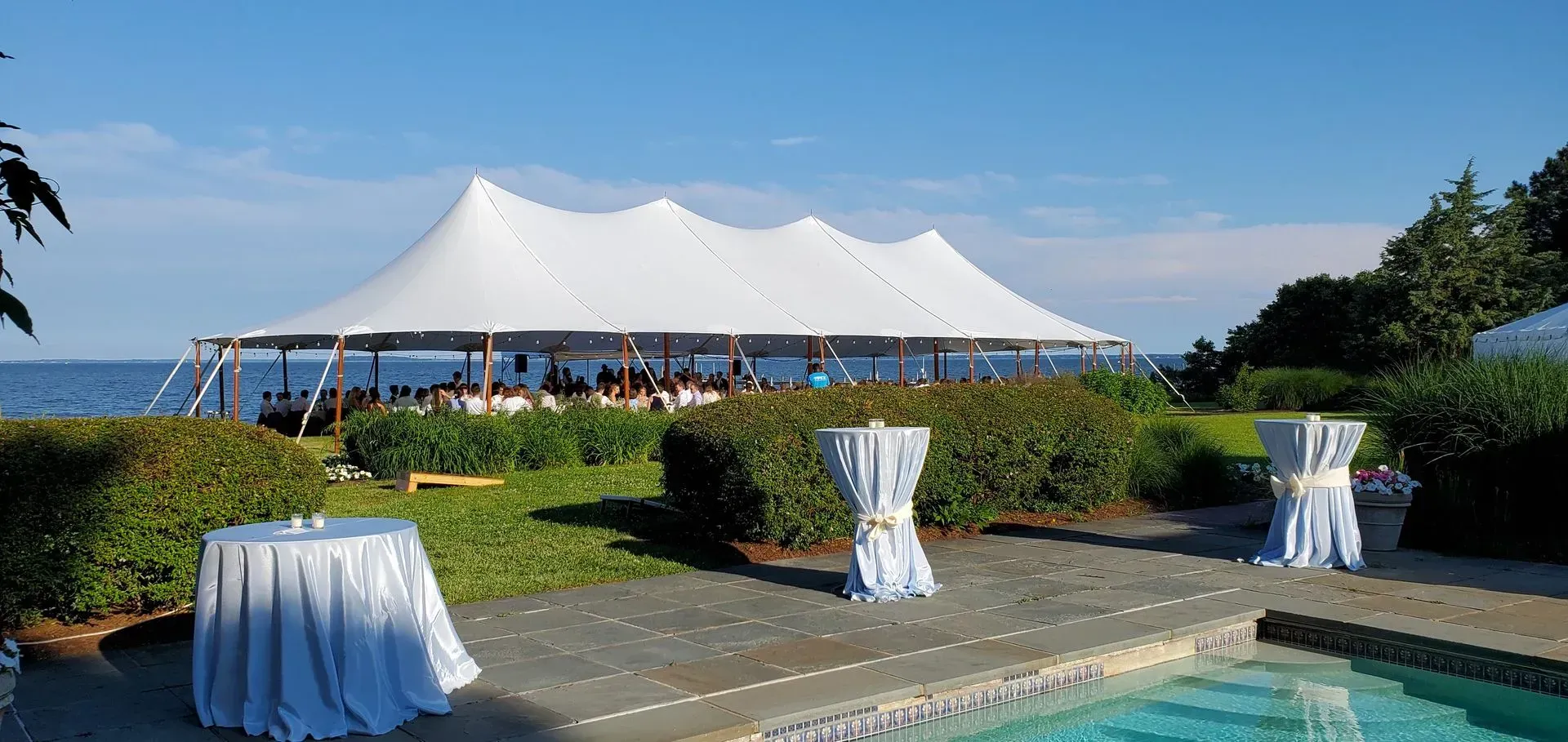 A white, multi-peaked event tent stands on a grassy lawn by the ocean, with draped high-top tables by a swimming pool.