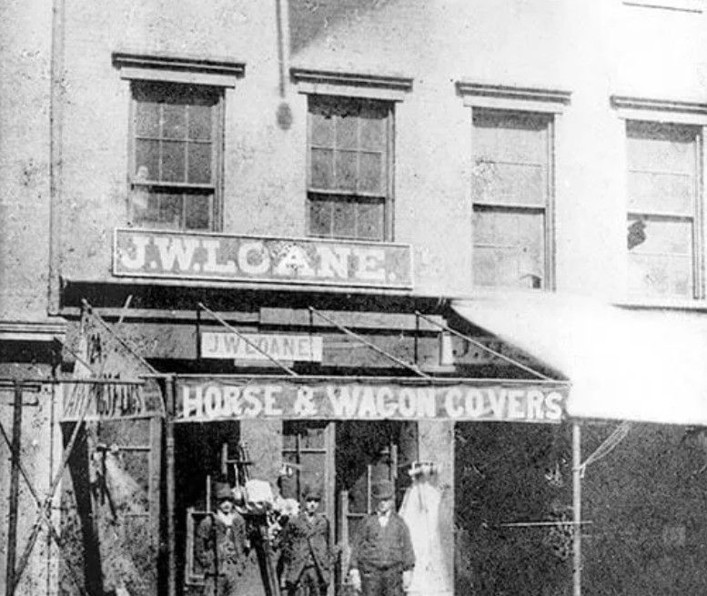A black and white photograph of J.W. Loane’s store, specializing in horse and wagon covers, with three people standing front.