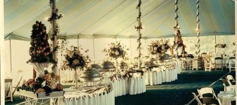 An elegant catering buffet inside a spacious, white-draped tent, featuring silver warming dishes and floral centerpieces.