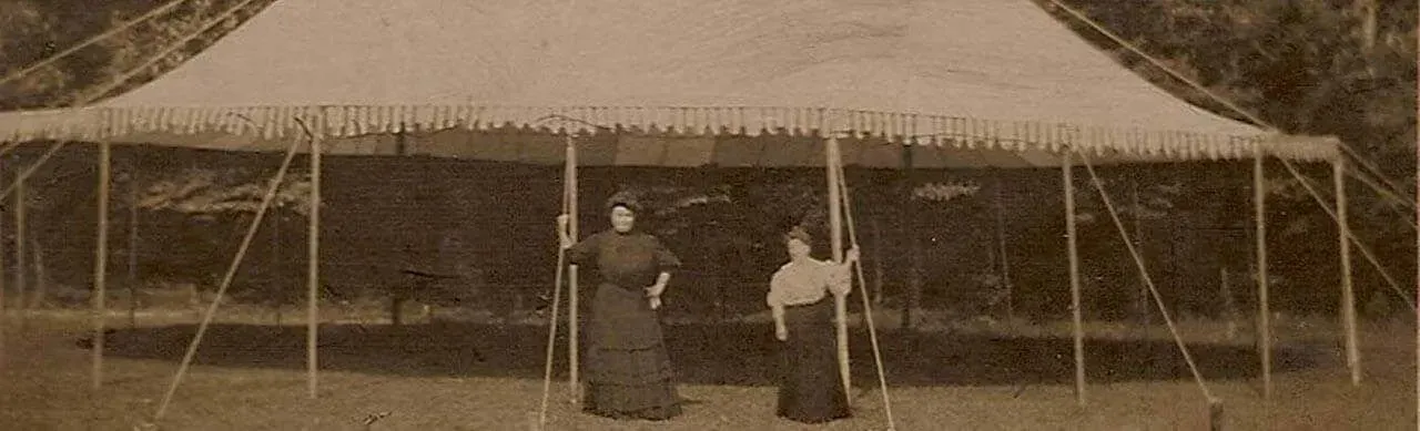 Two people in long, dark Victorian-era clothing stand side-by-side beneath a large, pale canvas tent in a grassy field.