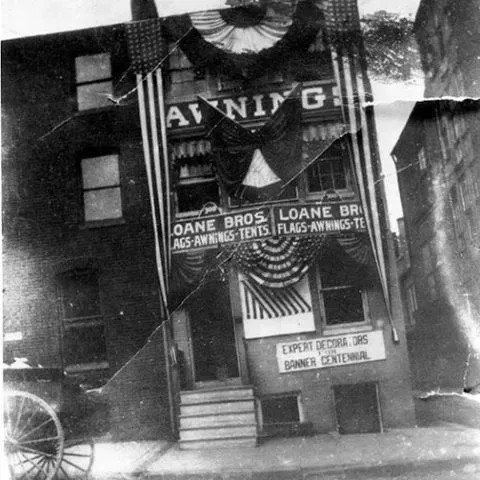 A historic brick storefront for Loane Bros. decorated with large American flags and bunting for a centennial celebration.