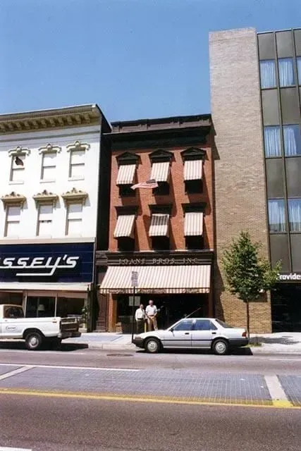 A narrow three-story brick building with striped awnings stands between a white building and a modern glass office tower.