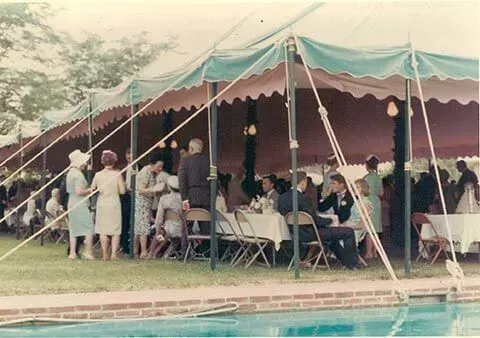 People at an outdoor garden party under a large green and pink tent next to a swimming pool.