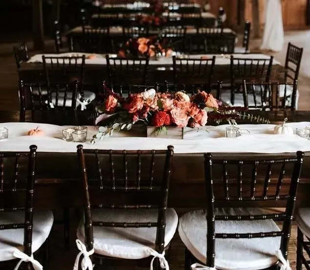 Long rustic tables set for a wedding, featuring white runners, dark wooden chairs, and lush fall-toned floral centerpieces.