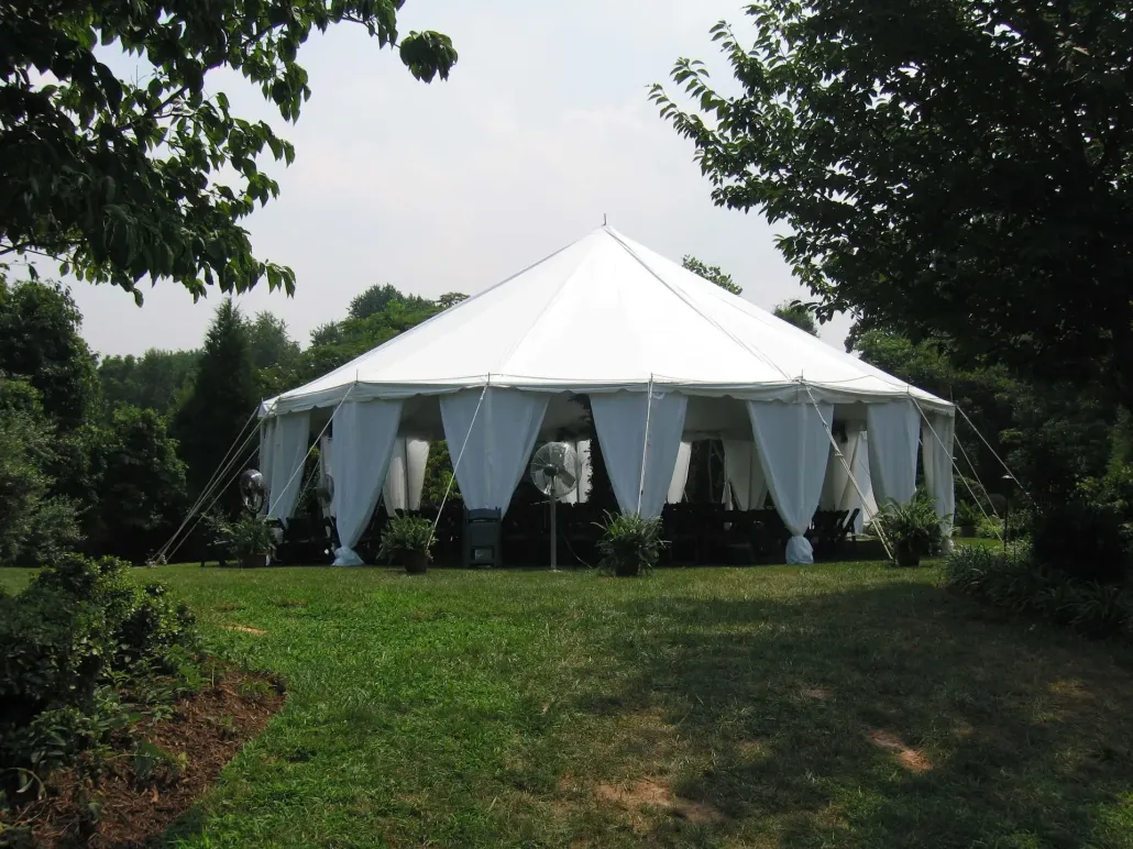 A large, white, octagonal event tent with draped curtains stands in a lush, green, wooded park.
