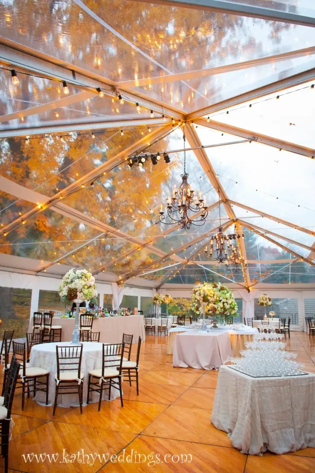 A wedding reception under a clear-roofed tent with chandeliers, wooden flooring, and tables decorated with floral centerpieces.