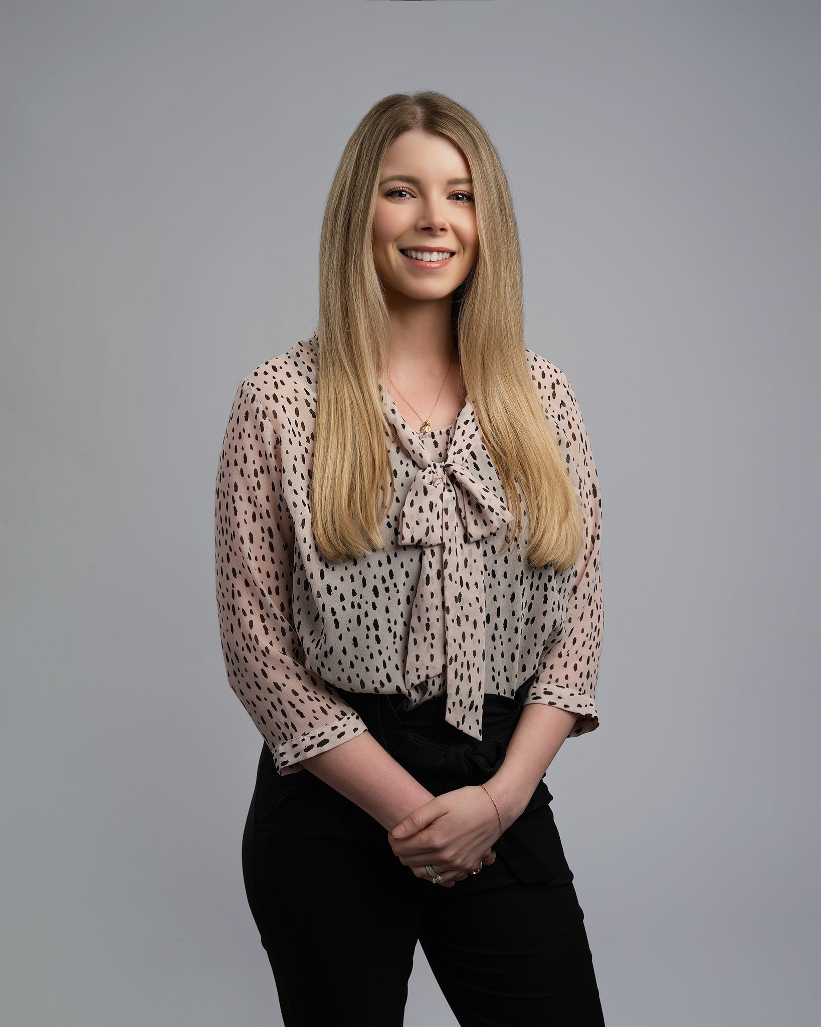 Smiling person standing against a gray backdrop, wearing a patterned blouse and black pants.