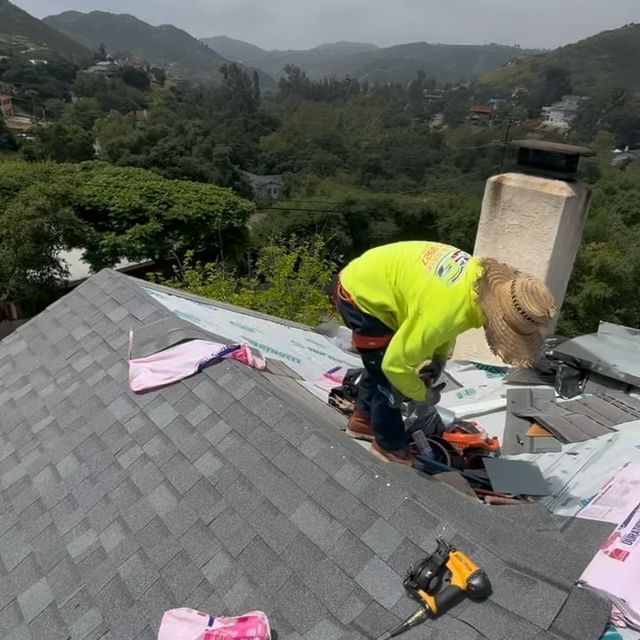 A man is working on the roof of a house with mountains in the background.