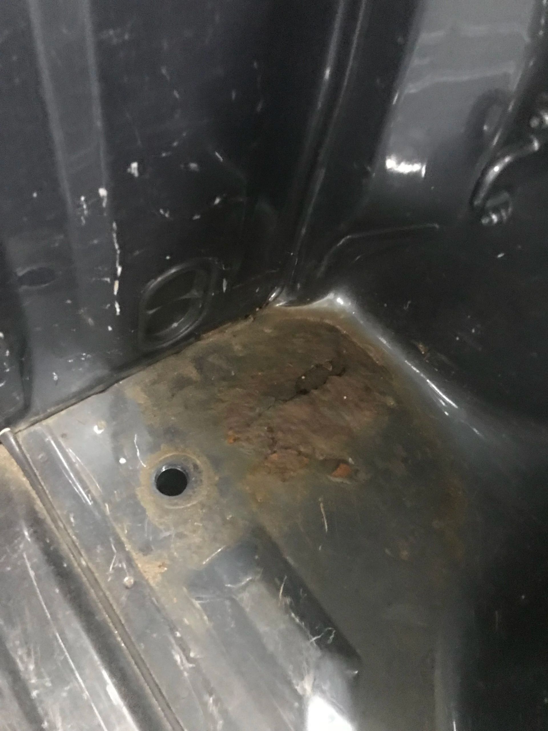 Rusty metal floor corner with a drain hole and worn black plastic panels