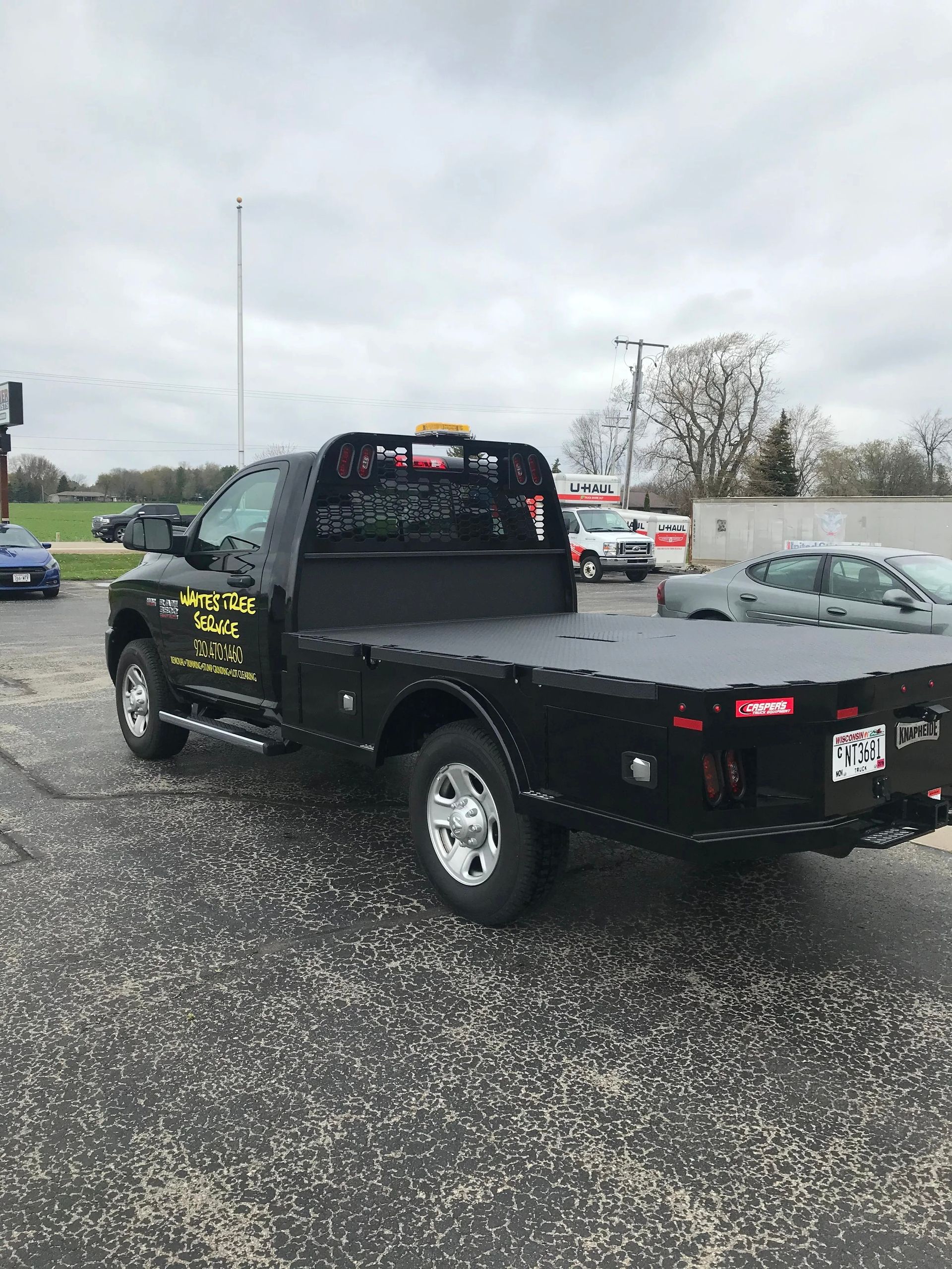 Black flatbed tow truck parked in a lot on a wet road, viewed from the rear side.