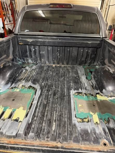 Empty pickup truck bed with worn black liner and exposed floor patches in a garage-like setting