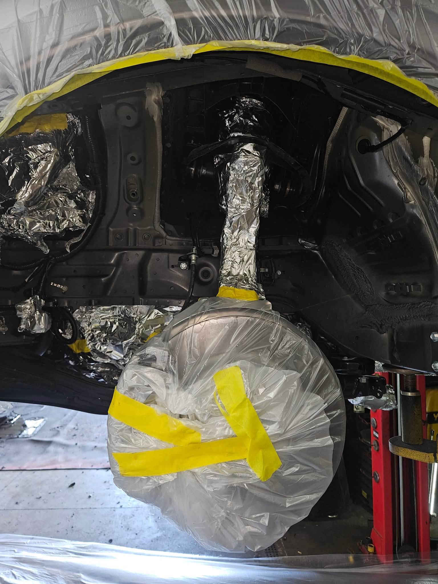 Car body in repair shop with rear wheel covered in plastic and yellow tape undercarriage visible