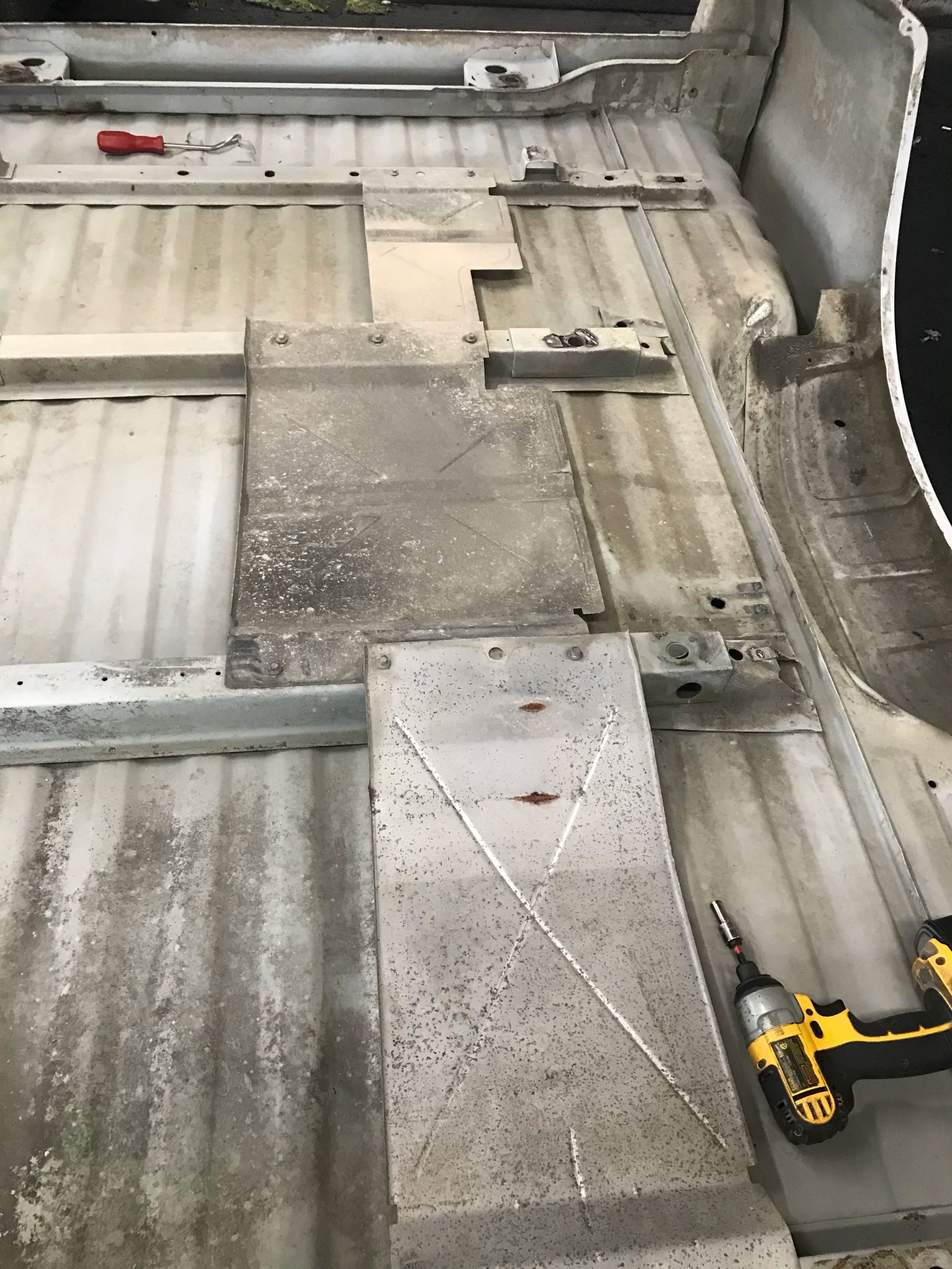 Car floor with exposed metal panels, scattered bolts, and a yellow cordless drill at bottom right.