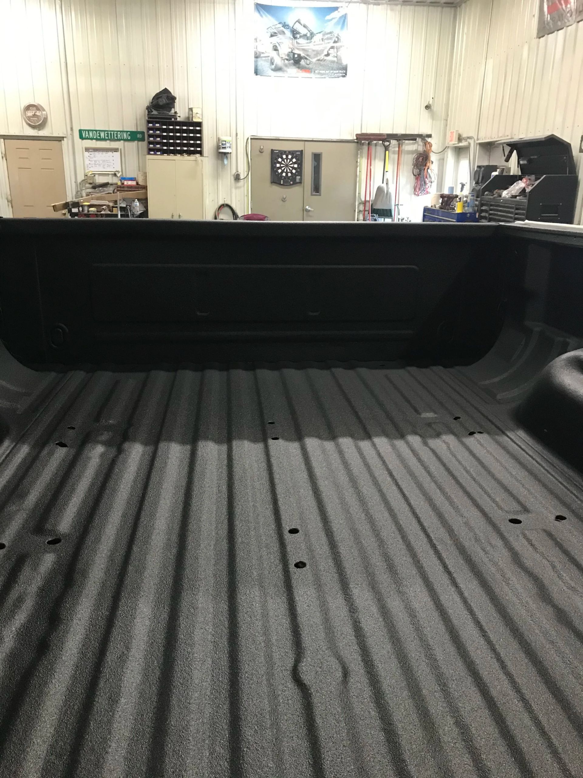 Empty pickup truck bed in a garage, with tools and equipment visible on the wall behind it.
