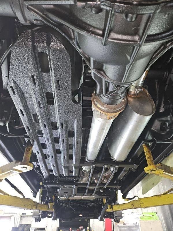 Undercarriage of a car on a lift, showing exhaust pipes and protective panels above a garage floor