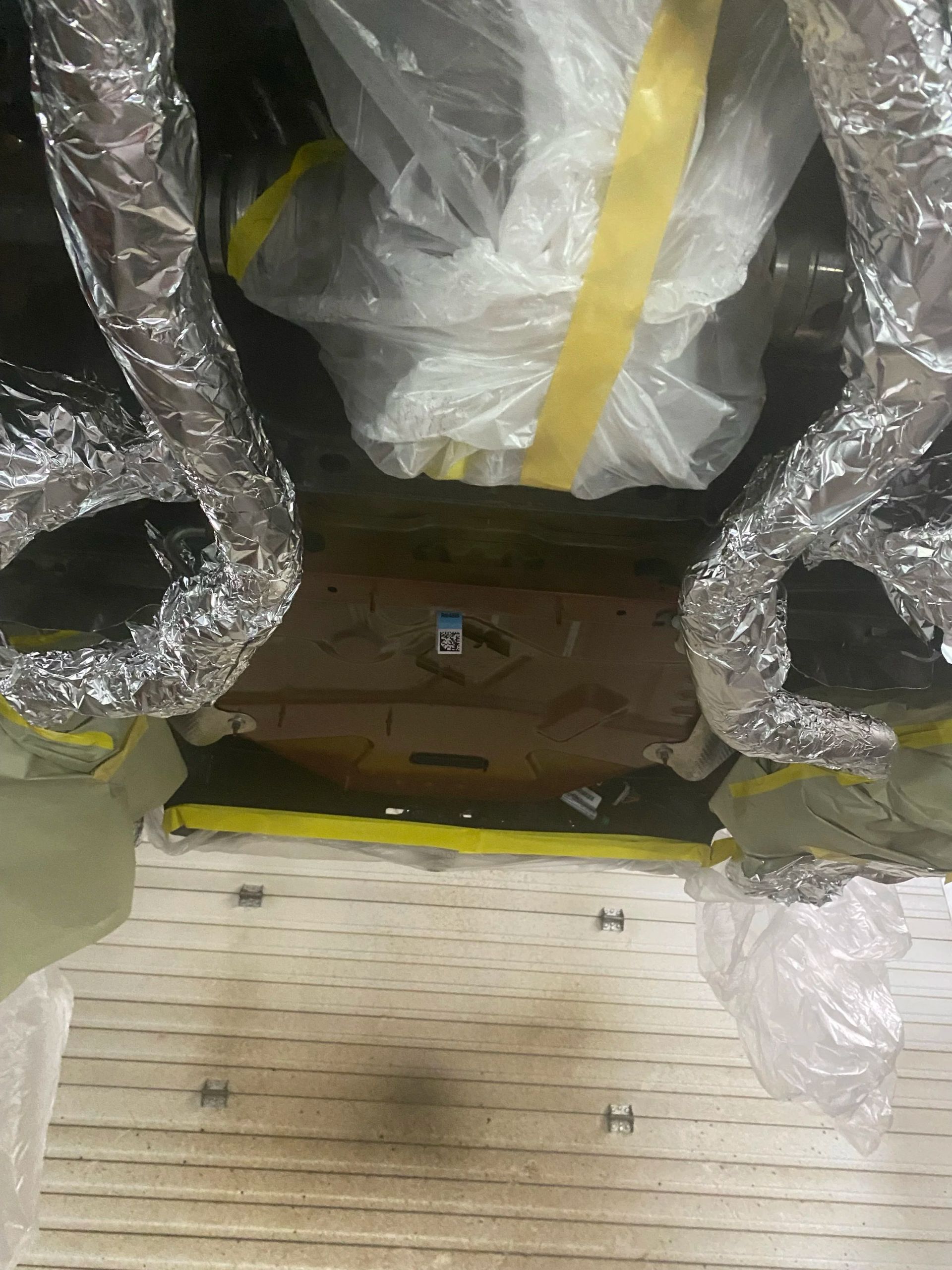 Plastic-wrapped machinery or furniture in a storage area, with yellow tape and a wooden floor visible underneath.