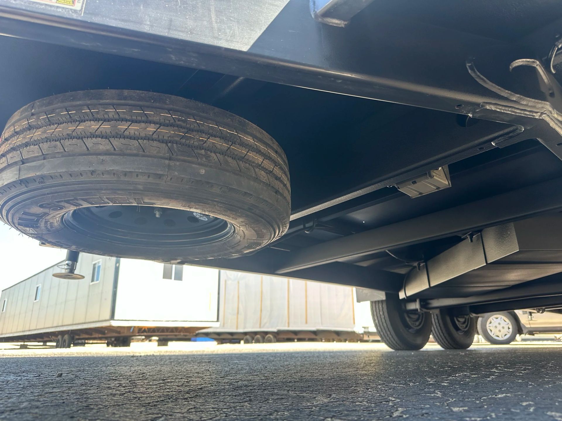Undercarriage of a trailer with spare tire and wheels, parked on gravel near shipping containers.