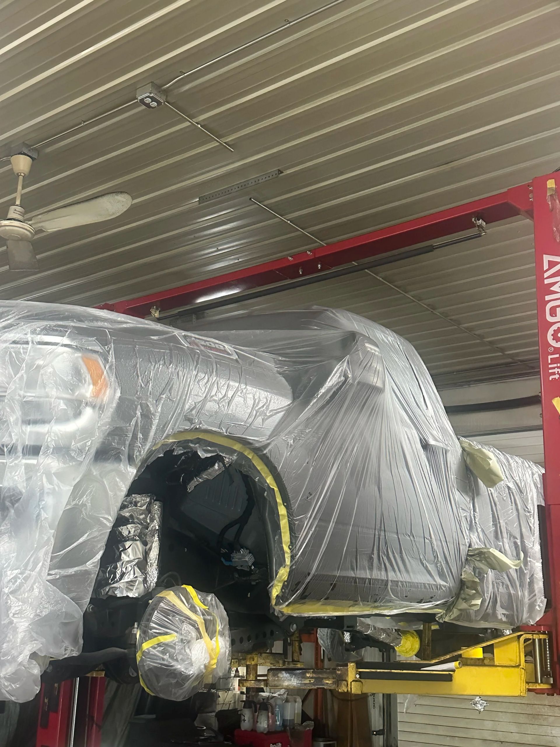Wrapped car body in a workshop, covered in clear plastic and primer, with a red lift post nearby