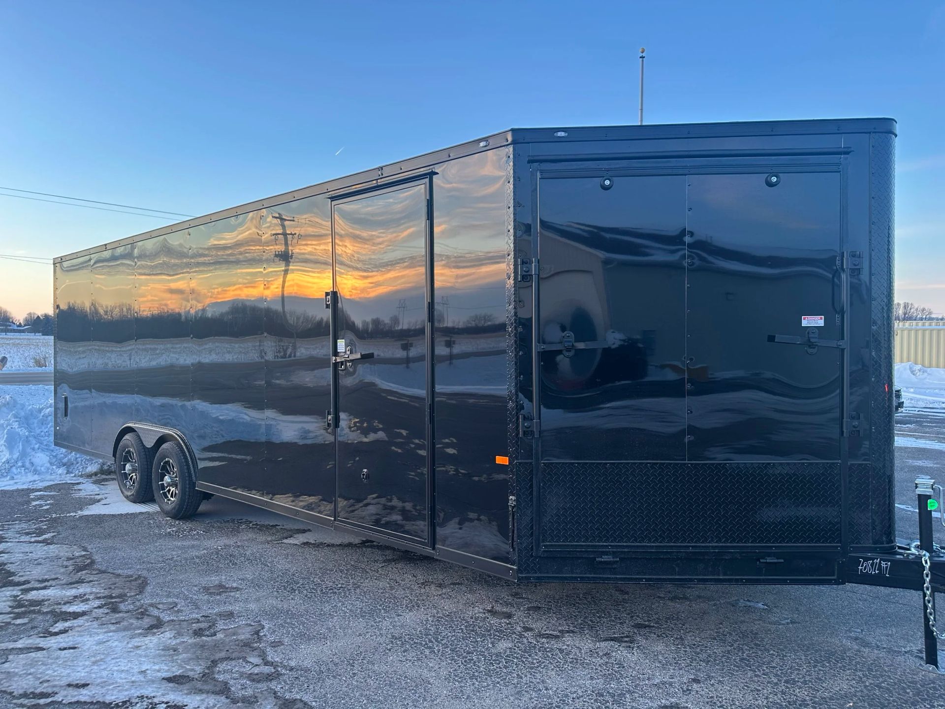 Black enclosed cargo trailer parked on snowy pavement at sunset, reflecting the sky.