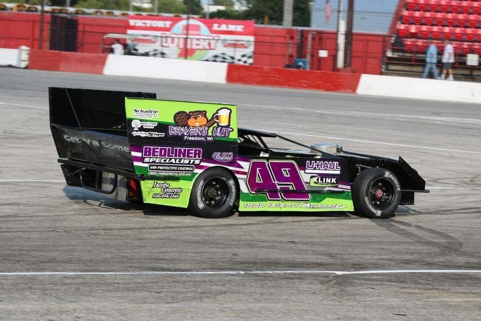 Black and green race car #49 speeding on a paved track with red barriers in the background
