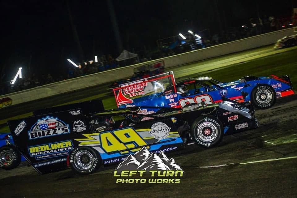 Two dirt track race cars, black #49 in front of blue and red car, speeding on a night track