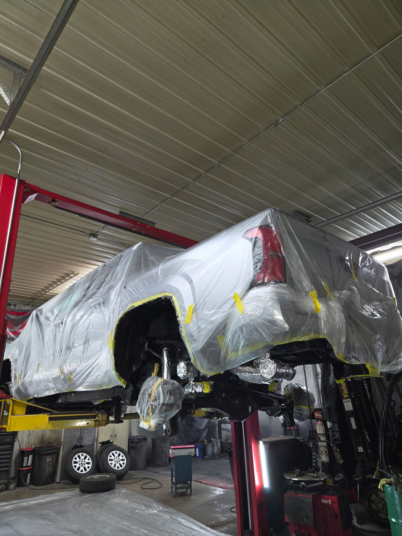 Car wrapped in plastic on a lift inside an auto repair shop, with wheels removed.
