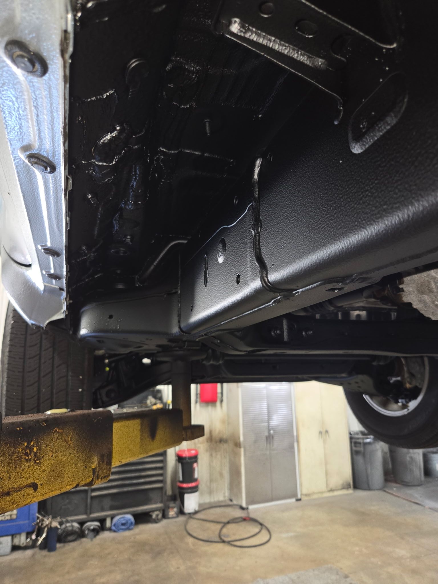 Undercarriage of a black car on a lift in an auto shop, with yellow lift arms visible.