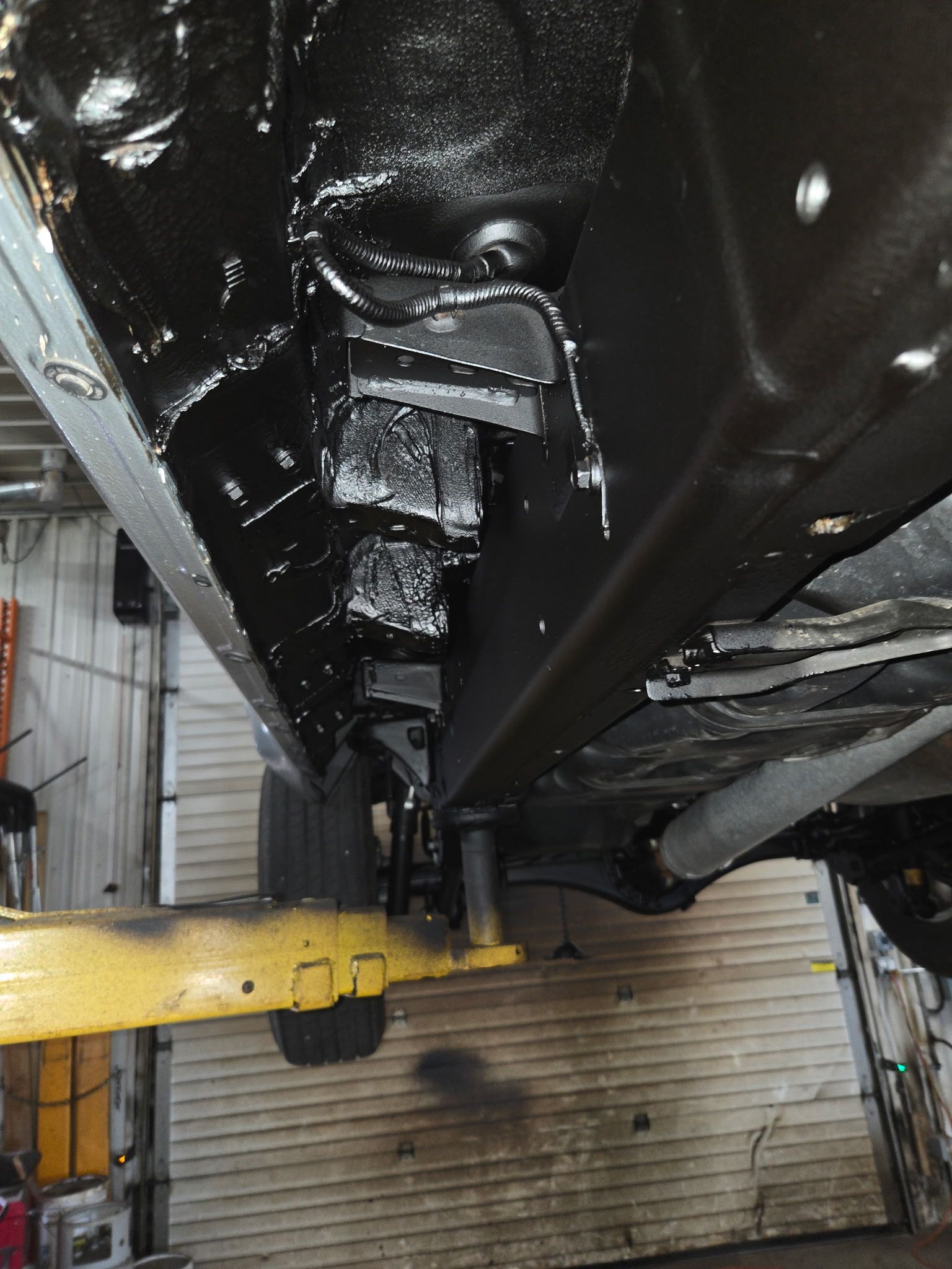 Undercarriage of a black vehicle on a lift in a garage, with a yellow support arm visible below.