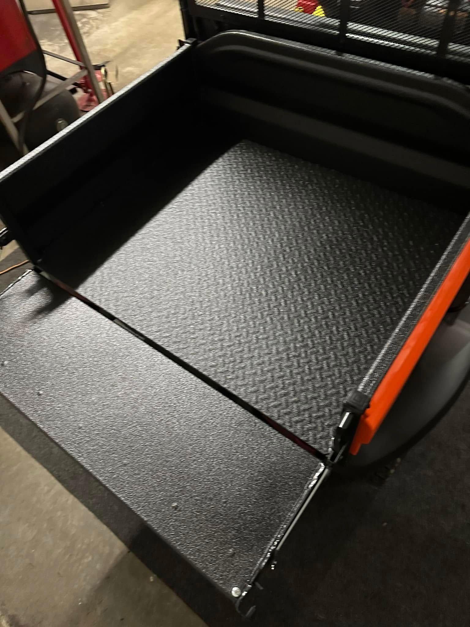 Black metal dump bed with textured floor and orange sides in a workshop.