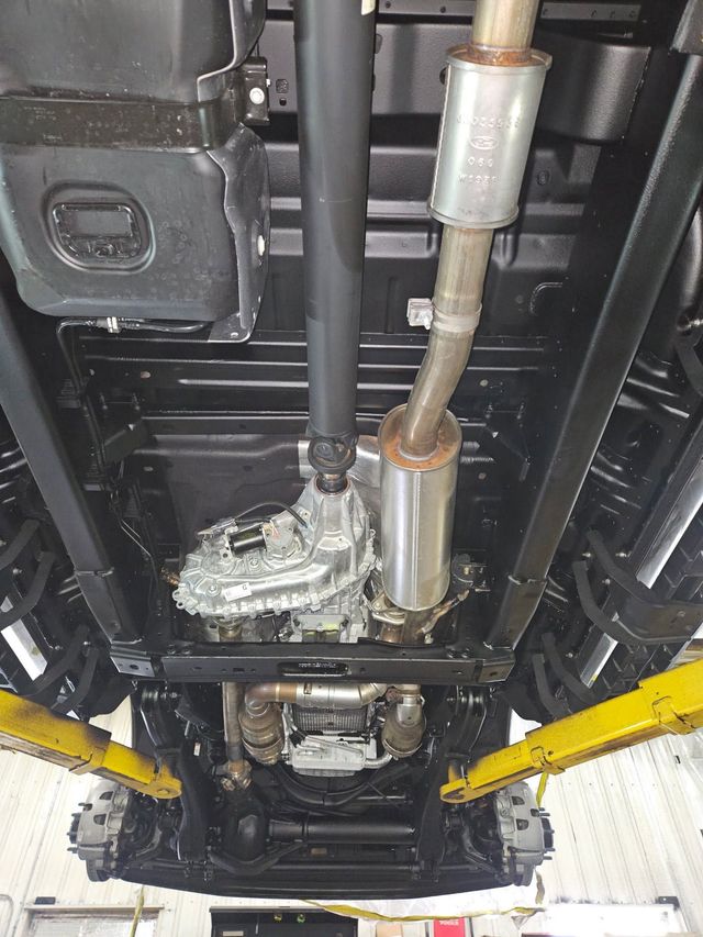 Underside of a car on a lift, showing exhaust pipes, suspension parts, and yellow lift arms.