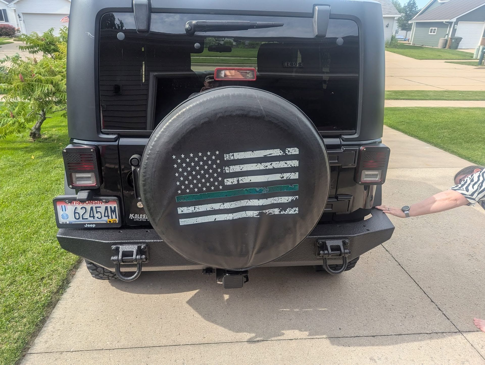 Black Jeep rear view with spare tire cover parked in a driveway, child’s arm reaching in from the right