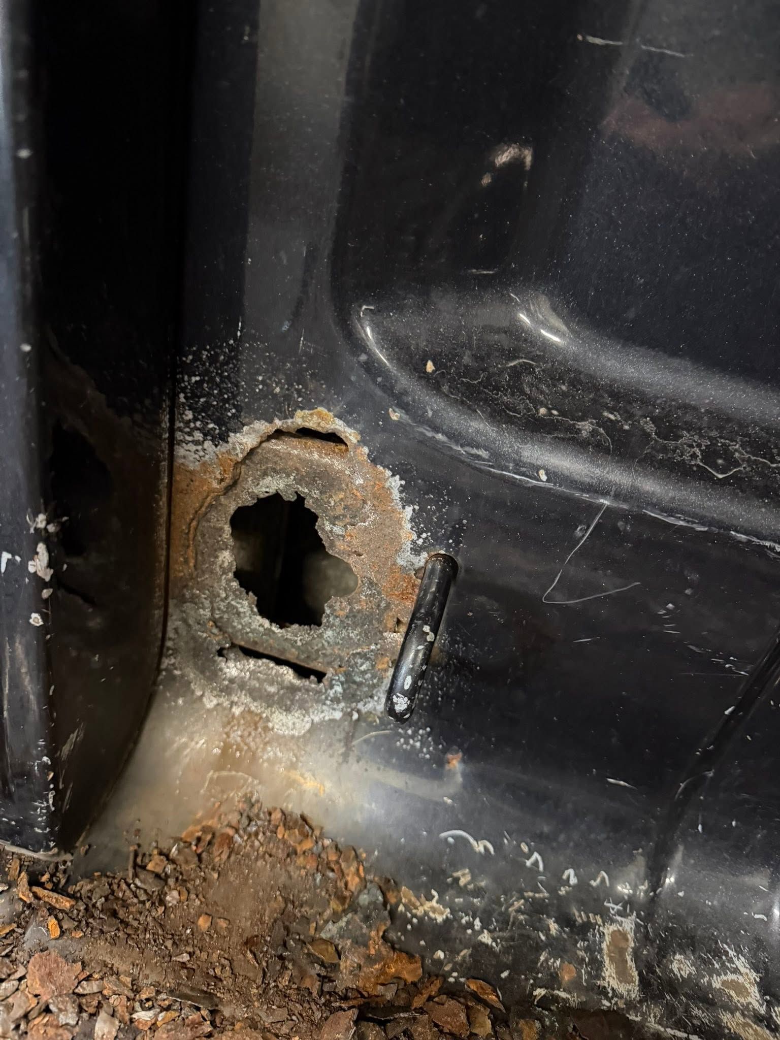 Close-up of a rusty hole in a black metal panel with dirt and corrosion around it.