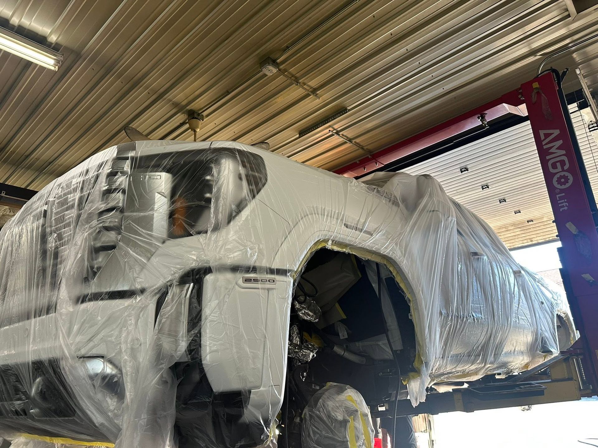Damaged white SUV wrapped in plastic inside an auto repair shop with a lift nearby