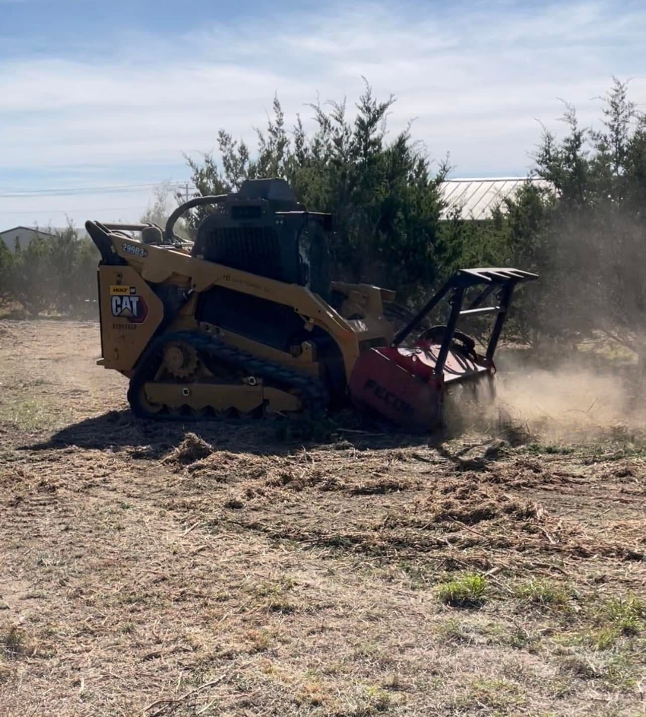 Yellow Caterpillar skid steer mulching brush on a dirt field; dust and debris in the air.