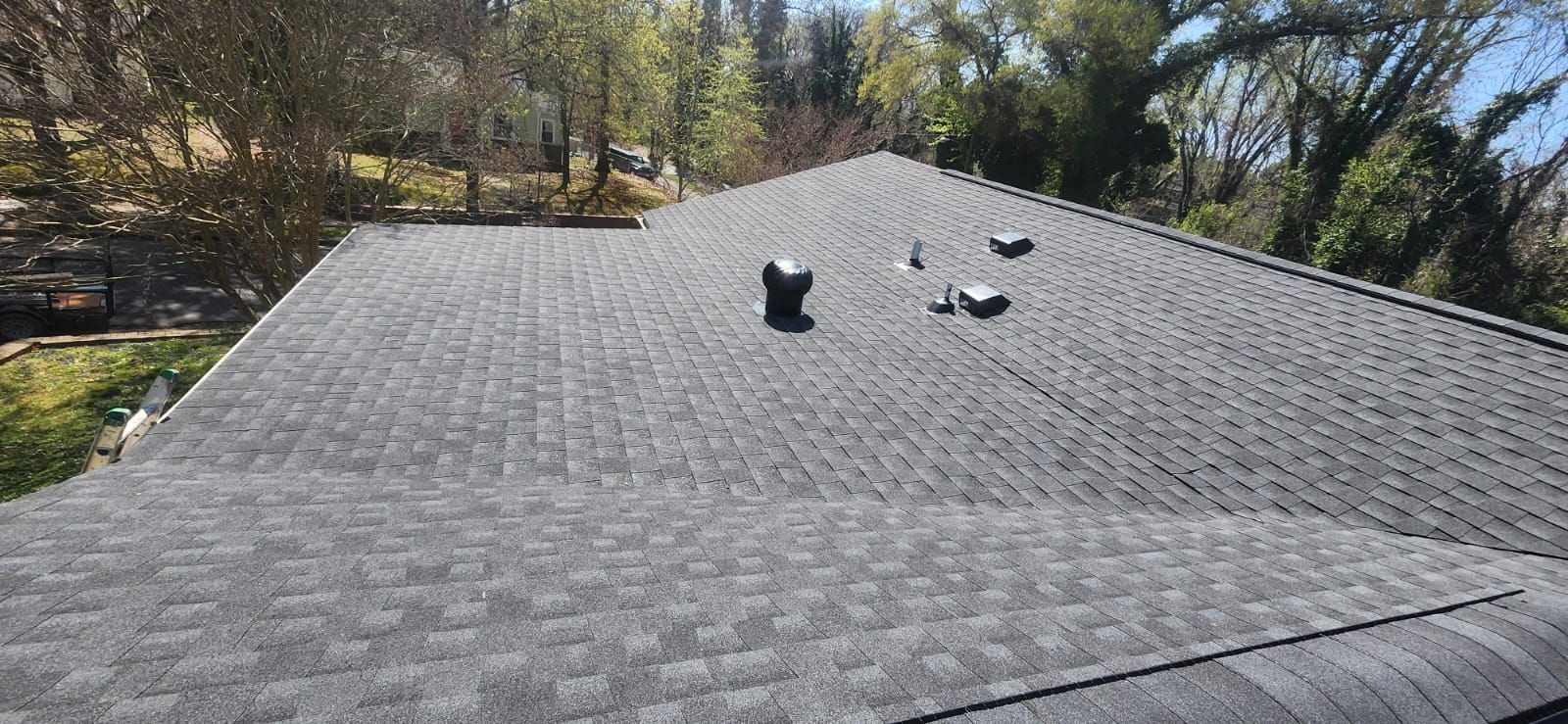 View of a dark gray shingle roof with vents, trees in the background. Sunny day.