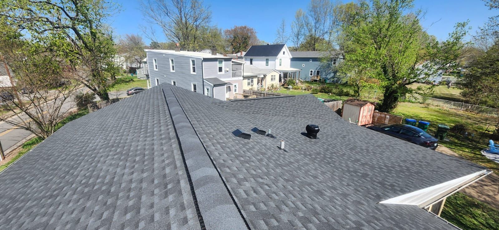 Overhead view of a gray shingled roof with a vent, houses, trees, and clear blue sky.
