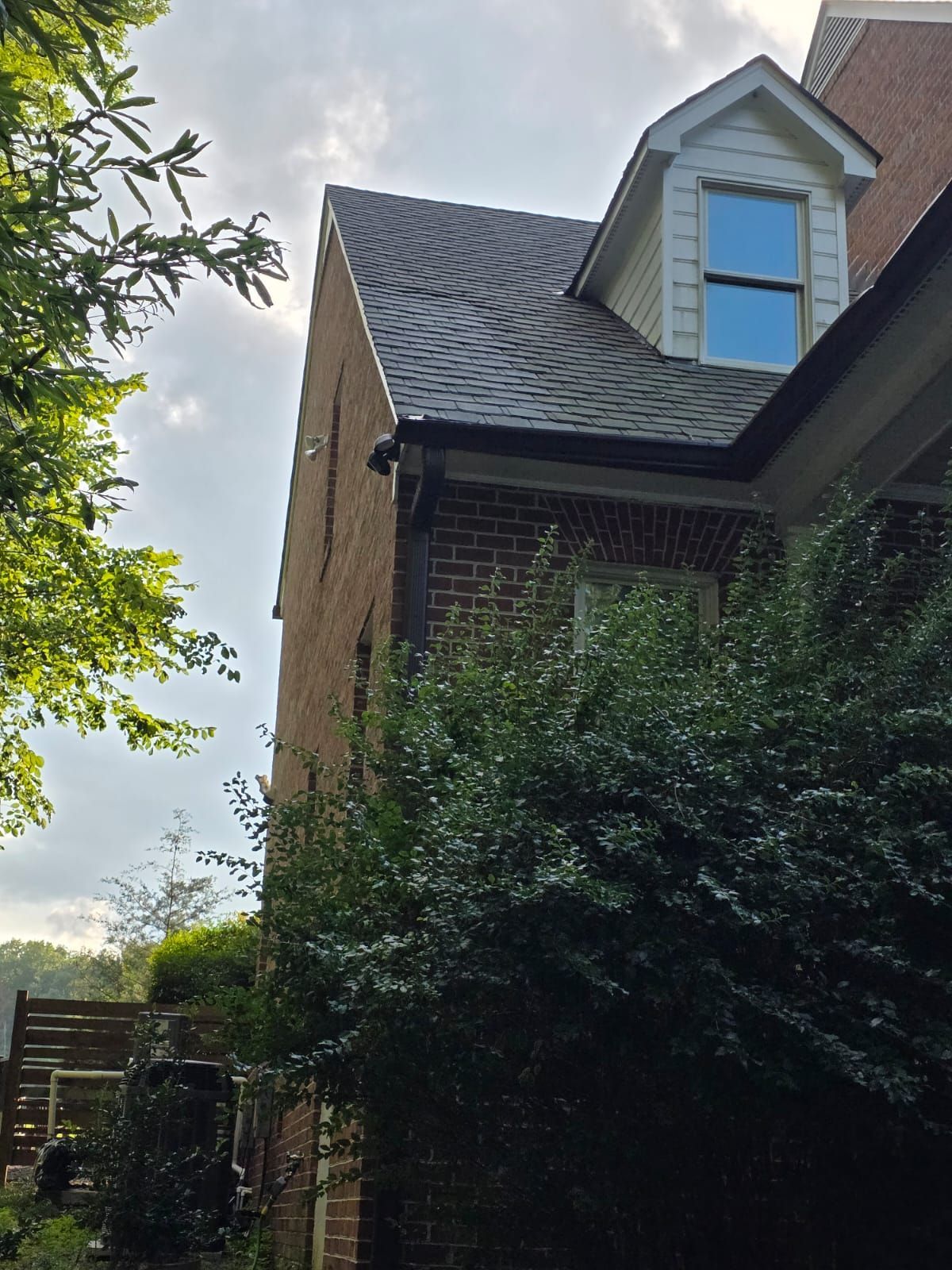 Brick building with dark gray roof, dormer window, and lush green bushes.