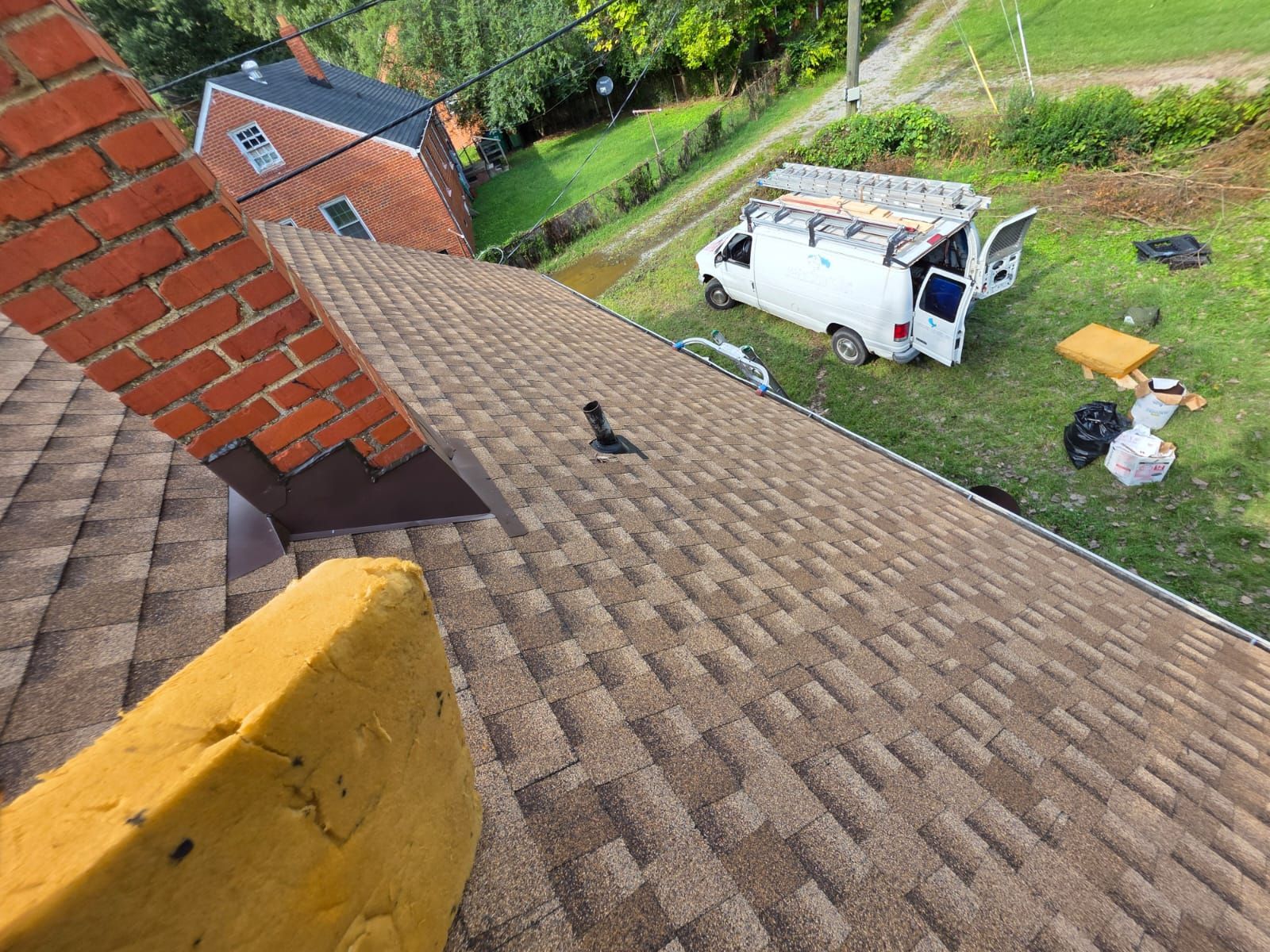 A brown shingled roof with a brick chimney and a white work van in the yard below.