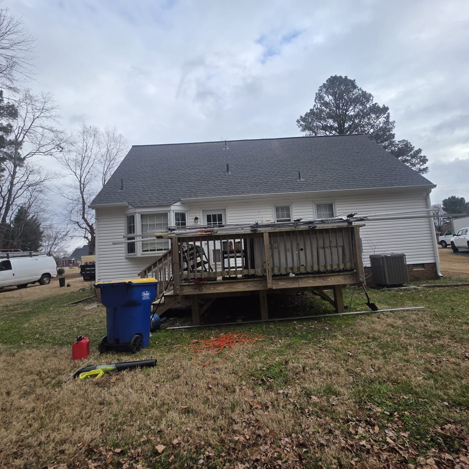 Backyard of a white house with a deck, blue trash can, and yard equipment on the grass under a cloudy sky.